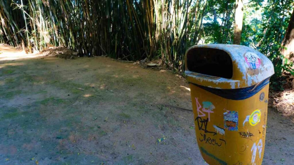 Indoor vs Outdoor Trash Cans: What's the Difference? A weathered outdoor trash can covered in graffiti stands along a dirt path surrounded by lush greenery.