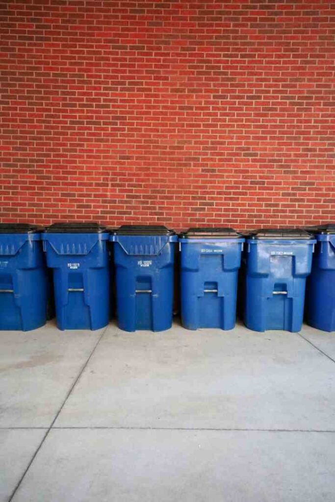 Indoor vs Outdoor Trash Cans: What's the Difference? A row of blue outdoor trash and recycling bins with black lids lined up against a red brick wall on a concrete surface.