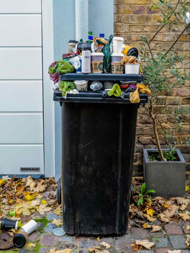 Indoor vs Outdoor Trash Cans: What's the Difference? An overflowing outdoor trash bin filled with takeaway cups, bottles, and food packaging beside a brick wall, with litter scattered on the ground.